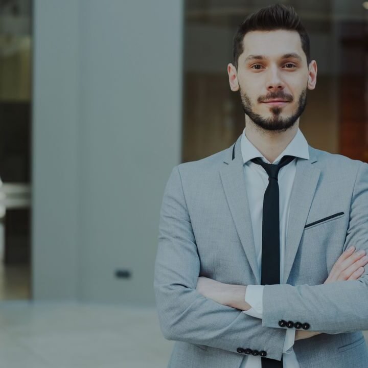 Businessman in a sleek gray suit with arms crossed, looking confident in a modern office.