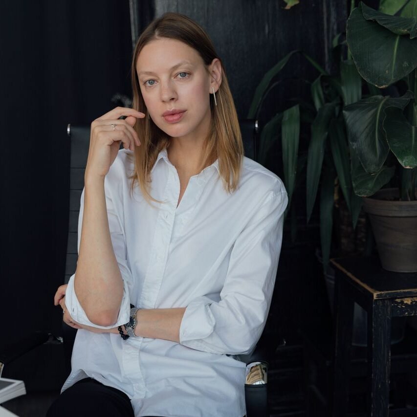 A stylish woman in a white shirt sits confidently in a modern, plant-filled office setting.